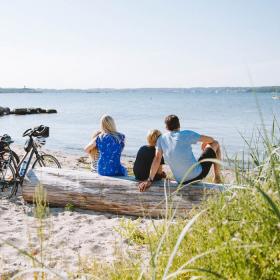 Familie sitzt mit Fahrrädern am Strand und schaut aufs Wasser 